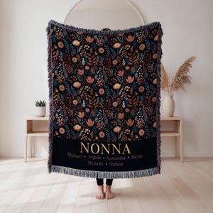 May include: A black woven throw blanket with a floral pattern in shades of orange, purple, and blue. The blanket has a black band with the name "NONNA" in gold lettering, along with several names below. The blanket has a fringed edge.