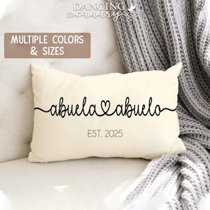 May include: Cream-colored rectangular pillow with the words "abuela & abuelo" in black script, connected by a heart symbol. The text "EST. 2025" is below. The pillow is on a white surface with a gray blanket in the background. The text "MULTIPLE COLORS & SIZES" is in a brown rectangle.