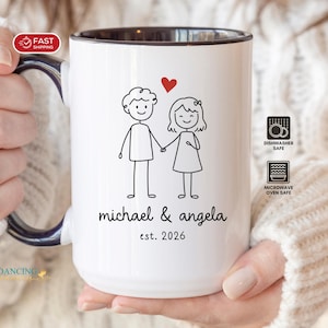 May include: White ceramic mug with a black rim and handle, featuring a stick figure couple holding hands, a red heart, and the names "michael & angela" with "est. 2026". The mug is dishwasher and microwave safe.