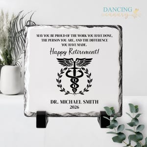 May include: A square, white stone plaque with a black border and a black stand. The plaque has the words "Happy Retirement!" and a medical symbol. The plaque also includes the name "DR. MICHAEL SMITH" and the year "2026".