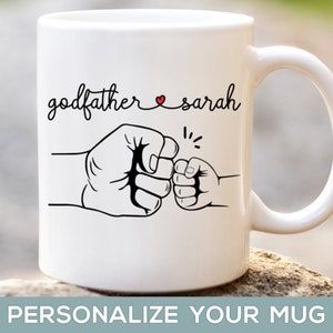 May include: White ceramic mug with a black and white illustration of two fists bumping. The text "godfather sarah" is written above the fists with a small red heart between the words.