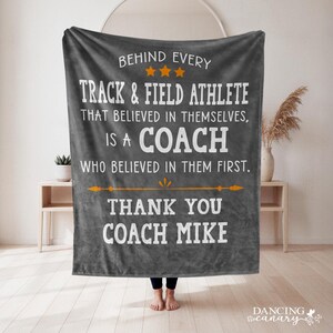 May include: A dark gray fleece blanket featuring white text that reads: BEHIND EVERY TRACK & FIELD ATHLETE THAT BELIEVED IN THEMSELVES, IS A COACH WHO BELIEVED IN THEM FIRST. THANK YOU COACH MIKE. Three orange stars and an orange line are included.