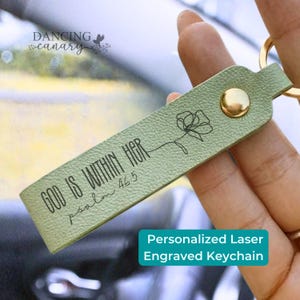 May include: A light green textured leather keychain featuring a gold metal snap button and a gold keyring. The keychain is laser engraved with a black line art flower and the text "GOD IS WITHIN HER psalm 46:5" in a script and sans-serif font.