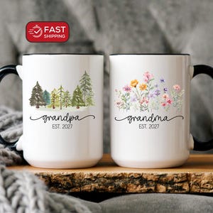 May include: Two white ceramic mugs with black handles and rims. The left mug features a forest graphic with the text "grandpa EST. 2027". The right mug displays a colorful wildflower bouquet graphic with the text "grandma EST. 2027".
