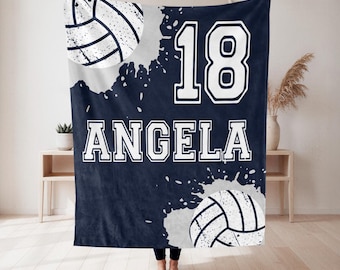 May include: A navy blue fleece blanket featuring a white volleyball graphic, the number 18, and the name ANGELA in a bold white collegiate font. The design includes white paint splatter accents on a dark blue background with a soft texture.