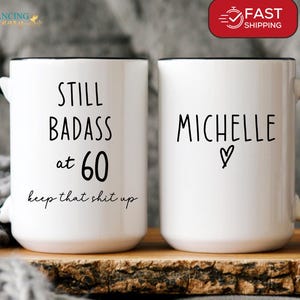 May include: Two white ceramic mugs with black handles and rims. One mug reads "STILL BADASS at 60 keep that shit up" in black text. The other mug has the name "Michelle" and a heart symbol. The mugs are on a wooden surface.