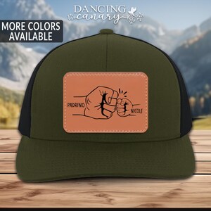 May include: Green and black trucker hat with a brown leather patch featuring two fists bumping. The patch says "Padrino" and "Nicole" on either side of the fists.