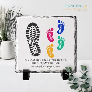 May include: A square, white stone plaque with a black border, featuring a black shoe print and colorful footprints. The text reads "STEPPED UP DAD" and "YOU MAY NOT HAVE GIVEN US LIFE, BUT LIFE GAVE US YOU. we love you."