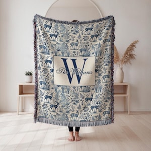 May include: A blue and white throw blanket with a deer and floral toile pattern. The blanket has a large navy blue initial "W" and the name "The Williams" in a rectangular frame. The edges are fringed.