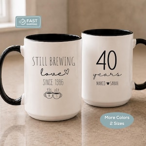 May include: Two white ceramic mugs with black handles and interiors. One mug features the text Still Brewing love since 1986 with a heart and two small coffee cups. The other mug displays the text 40 years and the names Marco and Sarah.