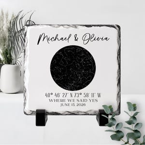 May include: A square slate plaque with rough, natural edges featuring a black circular star map in the center. The text reads Michael & Olivia, 40° 46' 27" N 73° 58' 11" W, WHERE WE SAID YES, and JUNE 15, 2026. It is supported by two black stands.