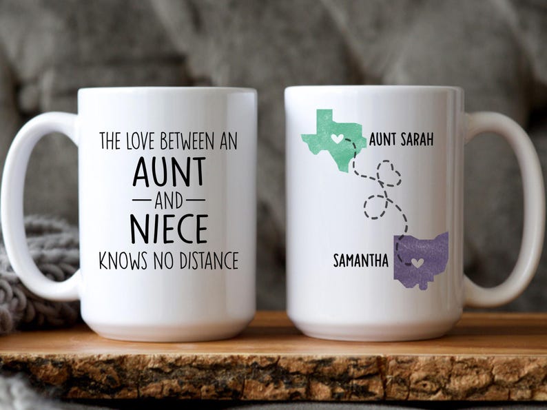 May include: Two white ceramic mugs with black text and a heart design. One mug says "The love between an aunt and niece knows no distance". The other mug has a map of Texas and Ohio with the names "Aunt Sarah" and "Samantha" and a heart design.