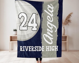 May include: A navy blue and grey fleece blanket featuring a large white volleyball graphic with the number 24, the name Angela in white script, and the text RIVERSIDE HIGH in white block letters on a vertical grey stripe background.