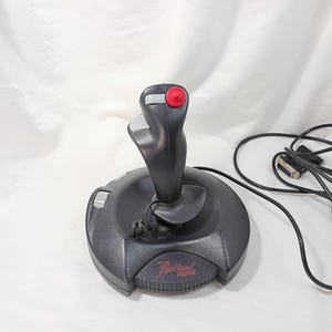 May include: A black gaming joystick featuring a red top button, a grey trigger, and a grey side dial. The base is circular with a textured grip area and displays the text Blackhawk digital in red lettering. A black cable with a connector is attached.