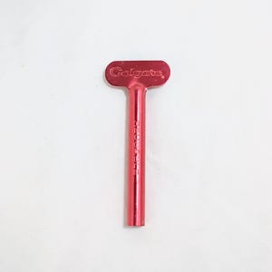 Peut inclure: Une clé de tube de dentifrice en métal rouge avec le nom de marque "Colgate" en relief sur la poignée. Le mot "RESCUE" est imprimé verticalement sur la tige de la clé. La clé est conçue pour presser le dentifrice.