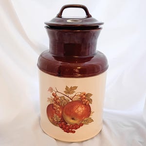 Pode incluir: Um pote de cerâmica vintage com tampa, com a parte superior marrom e a base creme. A base é decorada com uma ilustração colorida de maçãs e bagas. O pote tem uma alça na tampa.