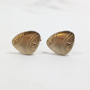 Puede incluir: Un par de gemelos dorados. Cada gemelo tiene una forma triangular redondeada con patrones decorativos grabados. El diseño incluye líneas curvas y un elemento en forma de estrella. Los gemelos están colocados sobre un fondo blanco.