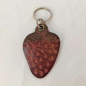 May include: A leather strawberry-shaped keyring with a silver key ring. The keyring is a deep reddish-brown colour with a textured surface, resembling the skin of a strawberry. The top of the keyring has a darker brown colour.
