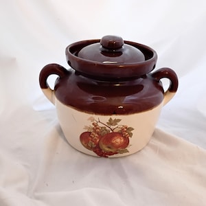 May include: A brown and cream-colored ceramic crock pot with a lid and handles. The crock pot features a fruit design with apples and grapes. The lid has a small knob. The crock pot is likely used for cooking or storage.