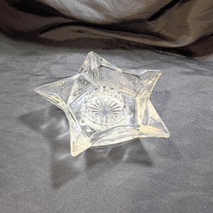 May include: A clear, star-shaped glass dish with a detailed, circular design in the centre. The dish has five points and is made of transparent glass, allowing light to pass through. The dish is likely used for decorative purposes.