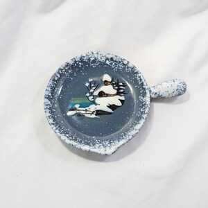 May include: A round ceramic spoon rest with a handle, featuring a dark blue base with a white speckled finish. The center displays a painted winter scene of a snow-covered house with dark windows and bare trees against a blue background.