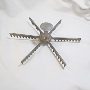 May include: A vintage, silver-toned cake cutter with six radiating arms, each featuring a serrated edge. The central hub has a rounded top and a cylindrical base. The tool is designed for evenly slicing cakes.