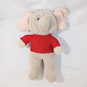 Puede incluir: Un peluche de elefante con cuerpo gris claro, orejas rosas y una camisa roja. El elefante tiene ojos azules y una boca rosa sonriente. El juguete es suave y tierno, ideal para niños.
