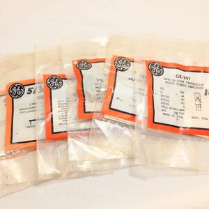 May include: A collection of five electronic components, each individually sealed in a clear plastic bag with an orange and white label. The labels feature the General Electric logo and technical specifications, including GE-247 NPN silicon transistor.