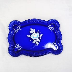 May include: A rectangular cobalt blue glass tray with a scalloped, ruffled edge. The centre features a hand-painted white floral motif with leaves, surrounded by four small white painted bows in the corners. The surface is smooth and glossy.