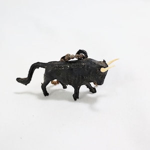 May include: A small, black bull figurine with tan horns and a decorative element on its back. The bull is posed in a walking stance, with its tail curved upwards. The figurine is set against a white background.