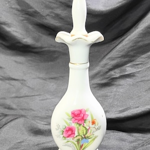 May include: A white porcelain perfume bottle with a floral design featuring pink roses and green leaves. The bottle has a long, slender neck and a decorative stopper. Gold trim accents the base and neck.