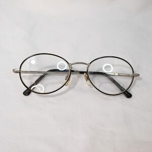 Könnte beinhalten: Ovale Brille mit schwarzen Rändern und silberfarbenen Metallrahmen. Die Gläser sind klar, und die Brille hat ein klassisches, schlichtes Design. Die Bügel sind dunkel, und die Nasenpads sind hell.