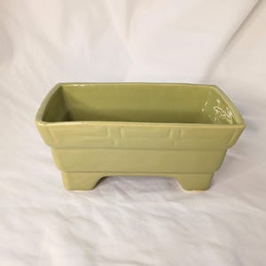 Pode incluir: Uma floreira retangular de cerâmica verde claro com um design texturizado na parte superior. A floreira é elevada sobre quatro pequenos pés. O interior está vazio, pronto para plantas ou outros itens decorativos.