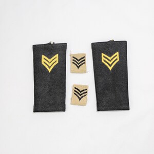 Puede incluir: Un conjunto de insignias de rango militar. Se muestran dos parches negros grandes con galones amarillos y dos parches beige más pequeños con galones negros sobre un fondo blanco. Los galones indican el rango.