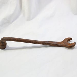 May include: A vintage, rusted metal wrench featuring a long, straight handle with an open-ended jaw on one side and a closed, cylindrical socket head on the opposite end. The tool displays a weathered, dark brown oxidized surface texture throughout.