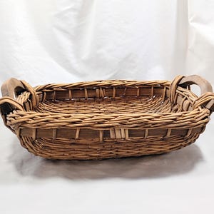 May include: A rectangular woven basket with wooden handles. The basket is made of light brown wicker and has a natural, rustic appearance. The handles are made of a lighter-colored wood and are attached to the sides of the basket.