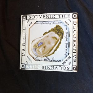 May include: Souvenir tile featuring an oyster design. The white tile has a gold and brown oyster illustration. Text includes "New Orleans", "Four Seasons of Louisiana", and "Souvenir Tile". The tile is framed with decorative text.