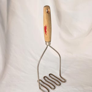 May include: A potato masher with a wooden handle and a stainless steel mashing head. The handle is light-colored wood with a red accent. The mashing head has a looped design.