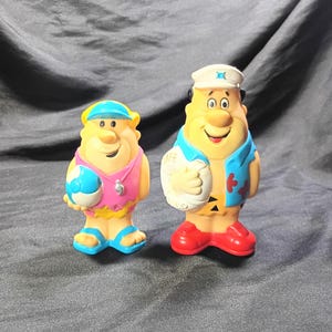 May include: Two plastic character figurines featuring a smaller figure in a pink tank top with a blue visor holding a beach ball, and a larger figure in a blue vest and white captain hat holding a white life preserver with the word LAKE printed on it.