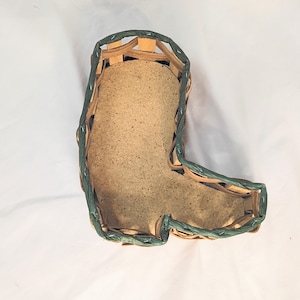 May include: A woven basket in the shape of a boot, with a light brown interior and a green woven border. The basket is made of natural materials and is likely used for storage or decoration. The boot-shaped design adds a unique touch.
