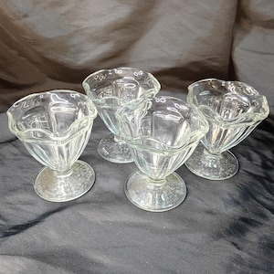 Puede incluir: Cuatro copas de helado de vidrio transparente con bordes festoneados y bases de pedestal. Las copas tienen un diseño clásico, perfectas para servir helado o postres. Las bases tienen un patrón decorativo.
