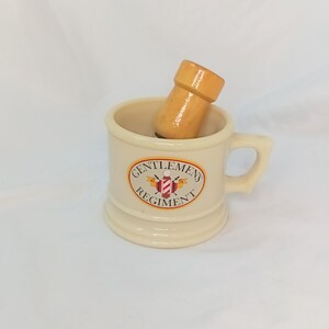 May include: A cream-coloured shaving mug with a handle and the words "GENTLEMENS REGIMENT" in an oval design. A wooden shaving brush is inside the mug. The mug has a classic, vintage aesthetic.