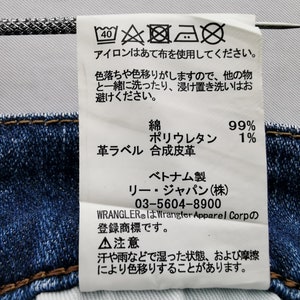 May include: A white tag with Japanese text and care symbols is attached to blue denim fabric. The tag includes washing instructions, fabric composition (99% cotton, 1% polyurethane), and brand information.