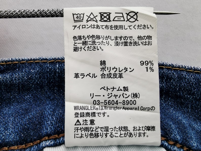 May include: A white tag with Japanese text and care symbols is attached to blue denim fabric. The tag includes washing instructions, fabric composition (99% cotton, 1% polyurethane), and brand information.
