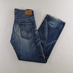 K&ouml;nnte beinhalten: Blaue Jeans aus Denim mit verwaschener Waschung und Used-Details. Die Jeans haben ein klassisches F&uuml;nf-Taschen-Design, ein rotes Levi's-Etikett und einen braunen Lederaufn&auml;her. Die Jeans sind gefaltet und liegen flach auf einer wei&szlig;en Oberfl&auml;che.