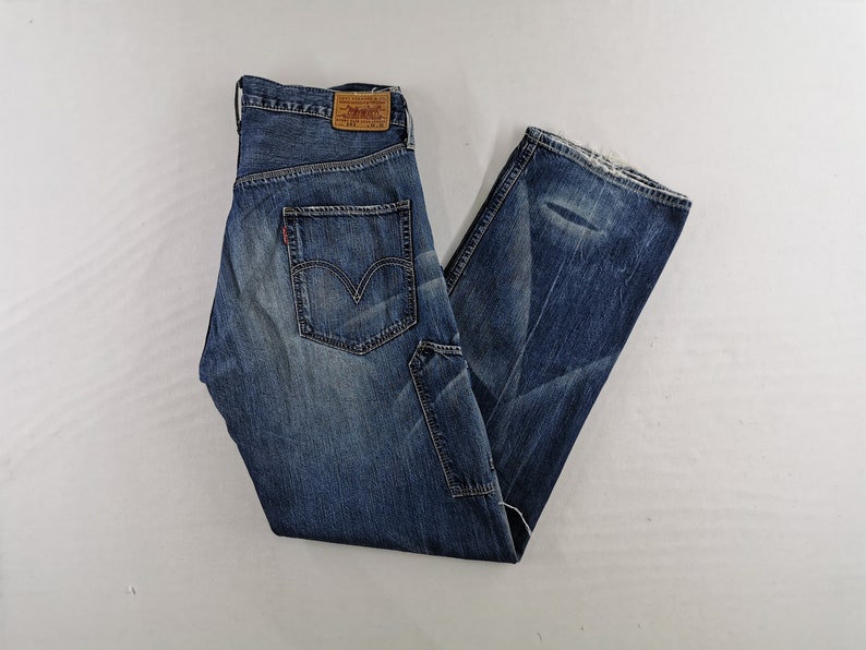 K&ouml;nnte beinhalten: Blaue Jeans aus Denim mit verwaschener Waschung und Used-Details. Die Jeans haben ein klassisches F&uuml;nf-Taschen-Design, ein rotes Levi's-Etikett und einen braunen Lederaufn&auml;her. Die Jeans sind gefaltet und liegen flach auf einer wei&szlig;en Oberfl&auml;che.