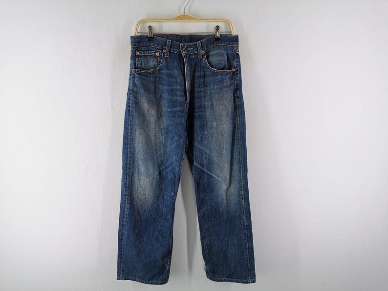 May include: Blue denim jeans with a faded wash and a straight leg fit. The jeans are hanging on a wooden hanger against a white background. The jeans have a classic five-pocket design.