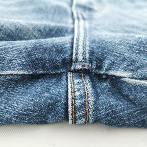 May include: Close-up of blue denim jeans fabric. The image highlights the seam and stitching details, with a focus on the texture and color variations of the denim. The stitching is in a contrasting yellow thread.