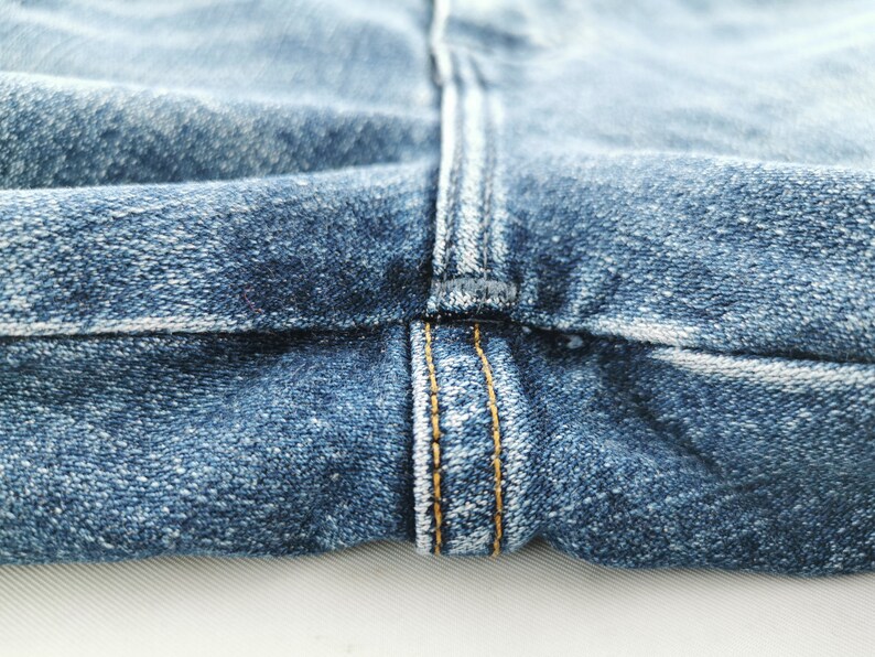 May include: Close-up of blue denim jeans fabric. The image highlights the seam and stitching details, with a focus on the texture and color variations of the denim. The stitching is in a contrasting yellow thread.