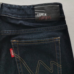 K&ouml;nnte beinhalten: Nahaufnahme einer dunkelblauen Jeans mit gelben N&auml;hten. Die Ges&auml;&szlig;tasche hat ein markantes 'W'-Nahtdesign. Ein rotes Edwin-Etikett ist auf der Tasche sichtbar, und ein schwarzes Lederpatch mit 'Edwin Jerseys' befindet sich am Bund.